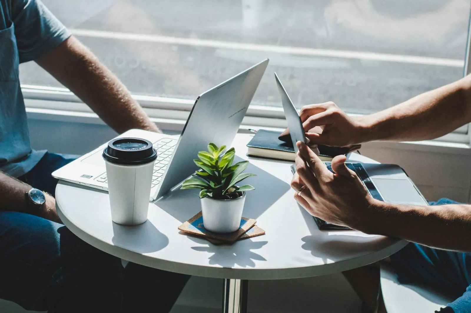 Two people at a small round table working on a laptop and tablet beside coffee and a potted plant
